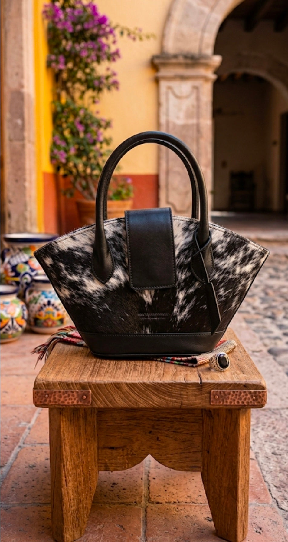 Black and cowhide leather flare tote bag on rustic wooden stool in Mexican courtyard